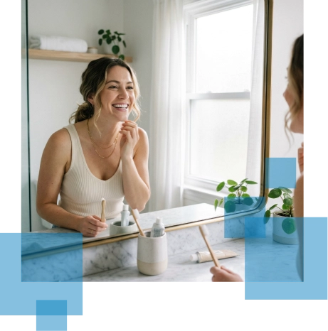 Smiling woman in a bathroom mirror applying skincare, surrounded by plants and natural light.