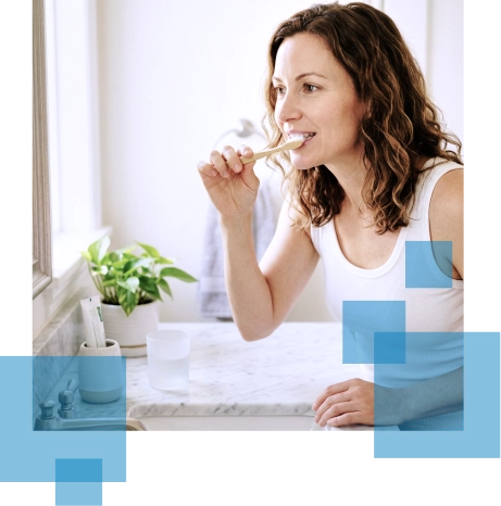 Woman brushing her teeth in a bright bathroom with a potted plant on the countertop.
