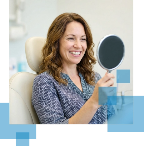Woman in a dental chair smiles while holding a mirror, reflecting her happy expression.